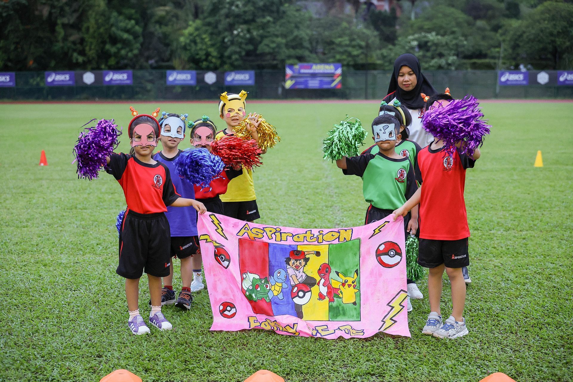 A group of children in costumes holding a colorful handmade Pokemon banner on a grassy field with an adult nearby.