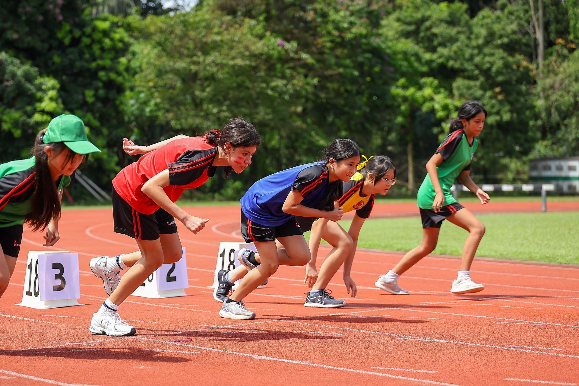Five runners in sportswear crouch at the starting blocks on a red track during a race.
