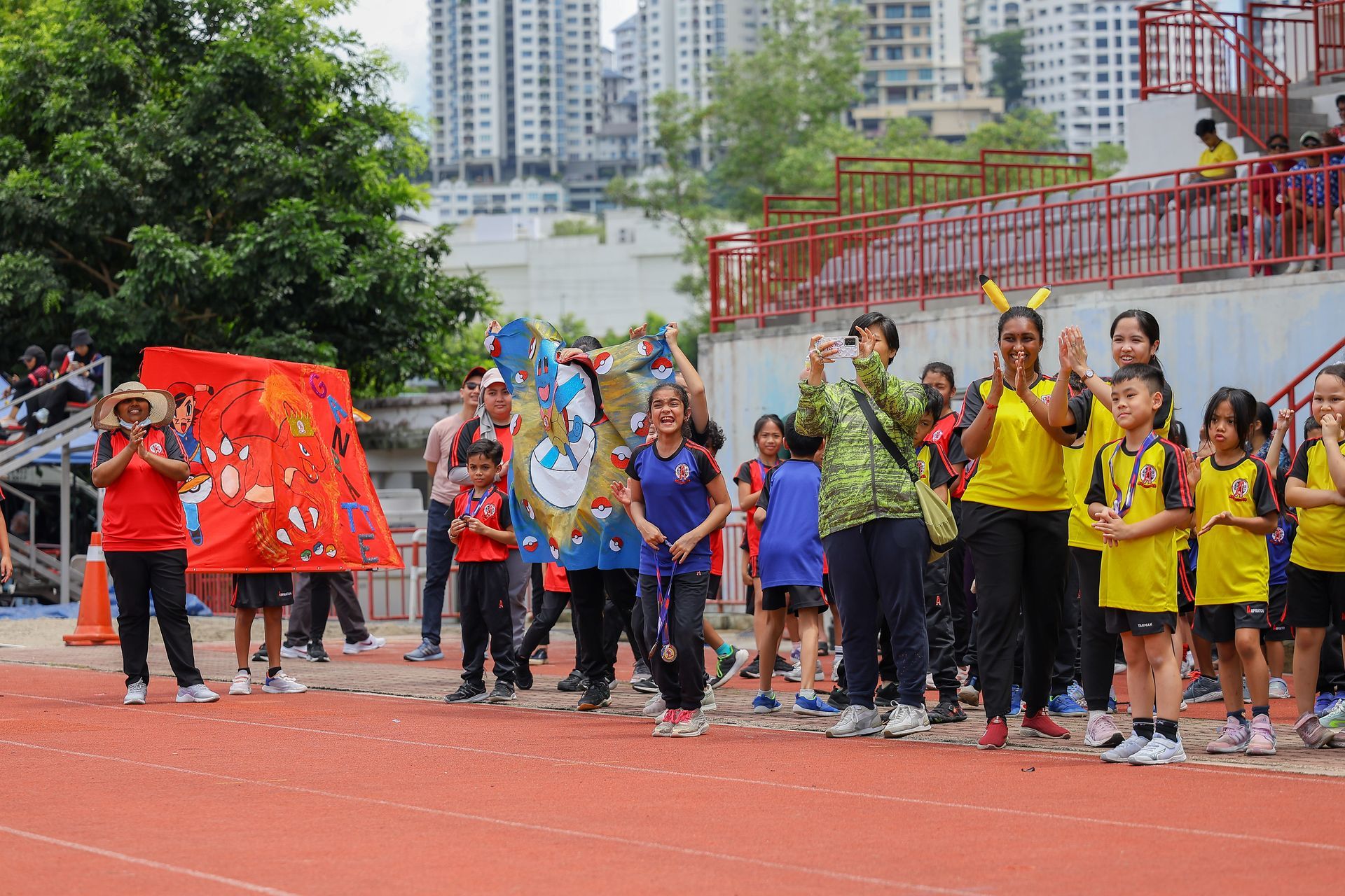 Students in colorful team uniforms and costumes cheer on a track field in front of a stadium.