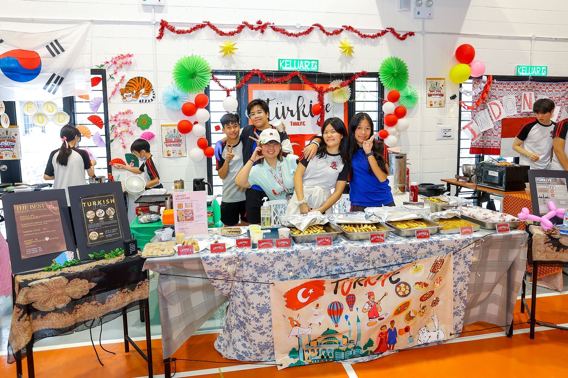 Students stand behind a decorated table with food and a Turkish flag at a school event, smiling at the camera.