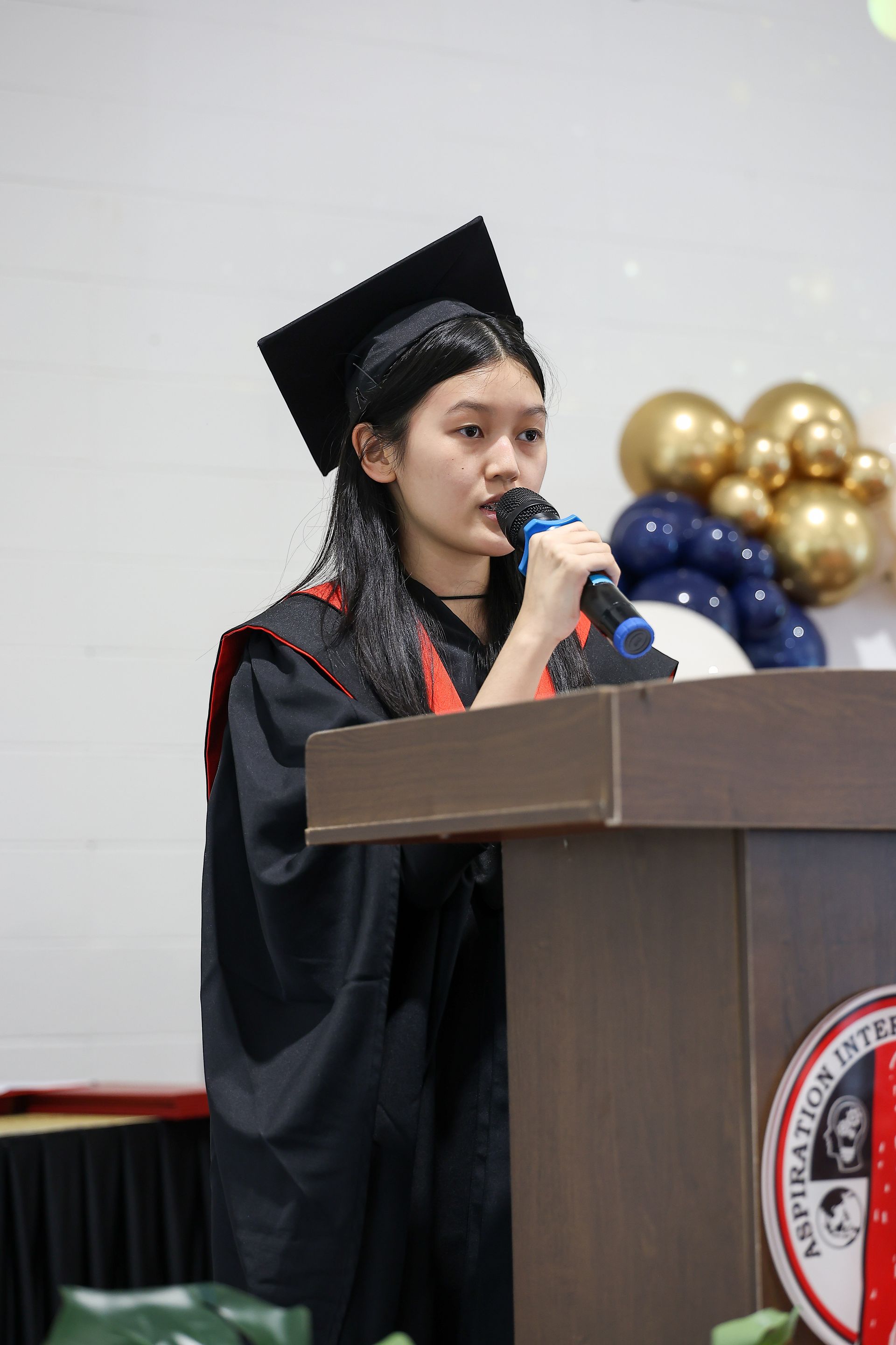 Students in graduation gowns and caps sit in a row during a ceremony with decorated balloons in the background.