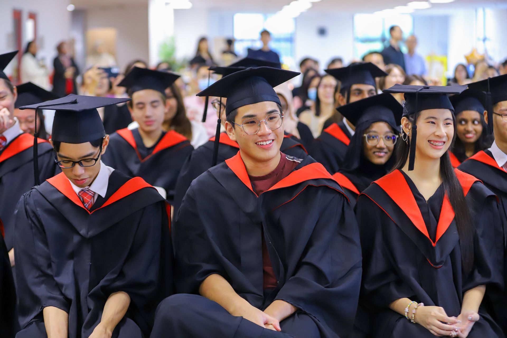 A group of graduates in black gowns and mortarboards sitting indoors, looking toward the camera with smiling expressions.