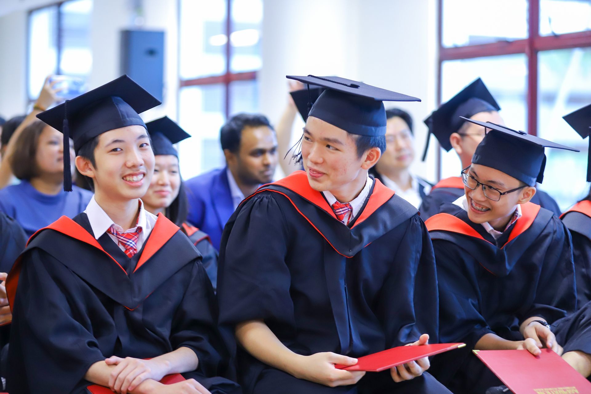 Graduates in caps and gowns sit together, smiling and holding red diplomas during an indoor ceremony.