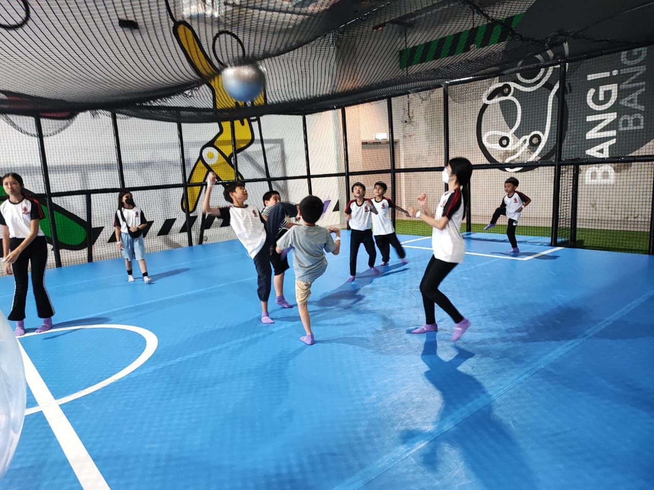Children in white shirts and black pants play with a large ball on a blue indoor court enclosed by black netting.