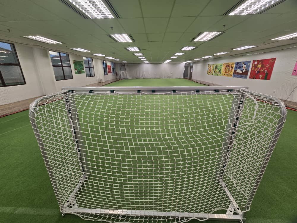 A small, indoor soccer goal on a green turf field, with ceiling lights and decorative wall art in the background.