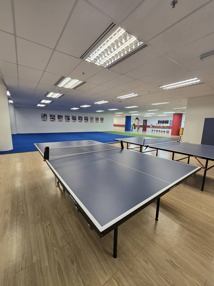 Two blue ping pong tables on a light wood floor inside a room with ceiling lights and a blue-matted area in the background.