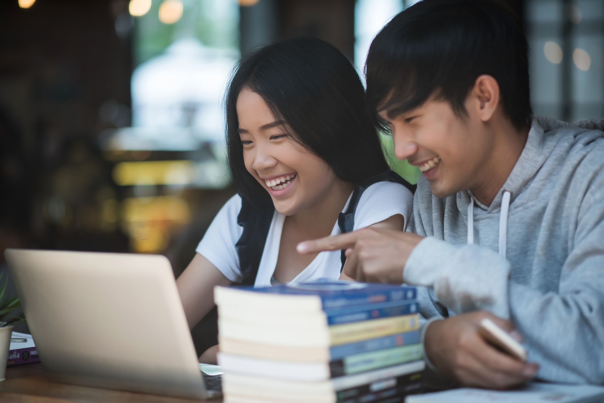 Two students sit at a table with books, laughing and looking at a laptop together.