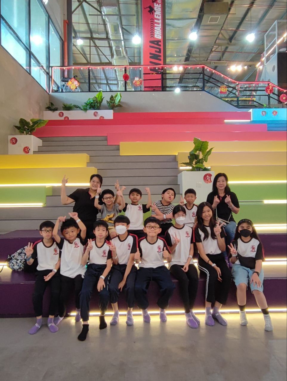 A group of students and teachers sitting on colorful tiered stairs inside a Ninja Park facility.