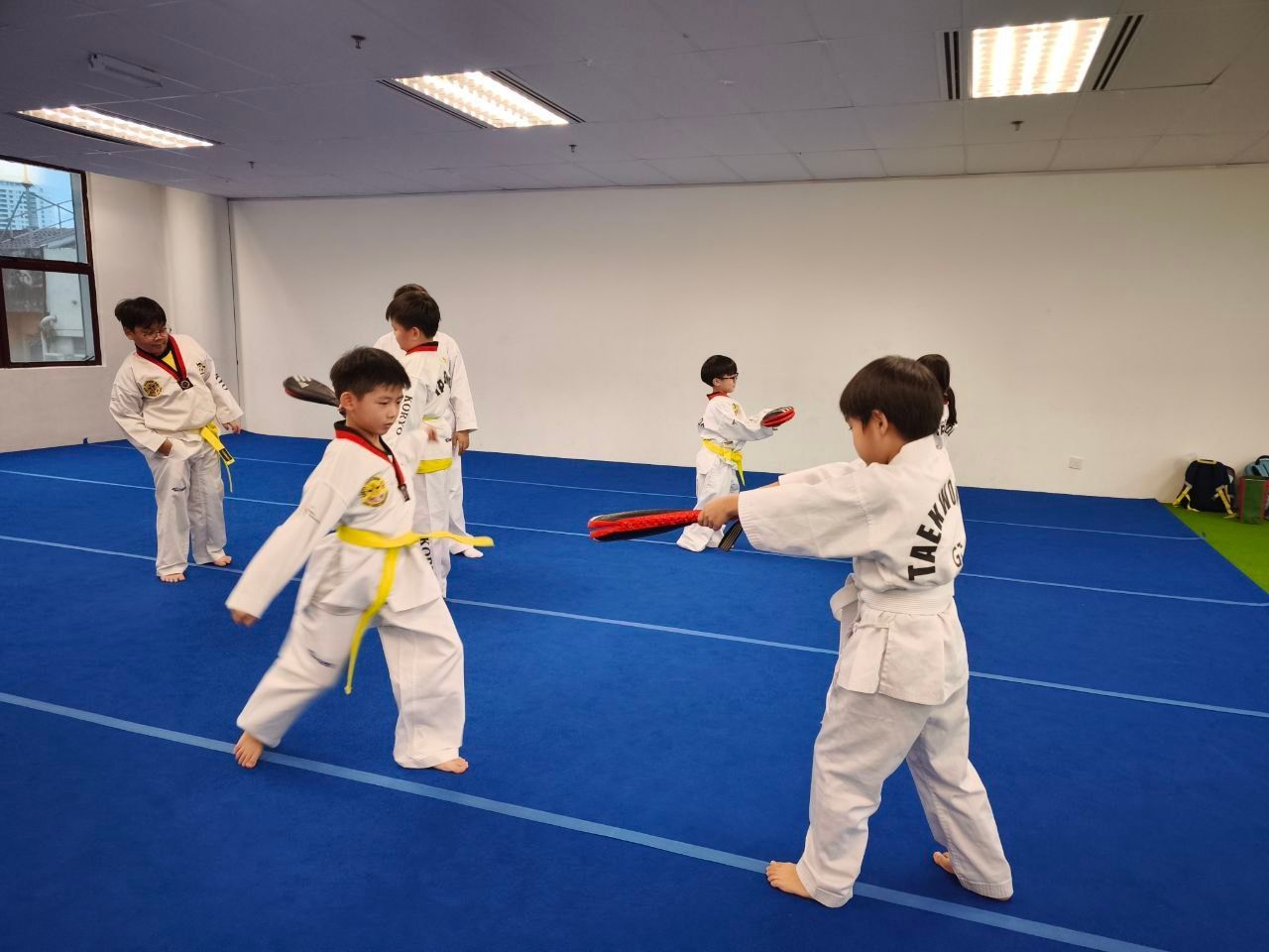 Children in white taekwondo uniforms practice kicking and striking drills on a blue matted floor in a training studio.