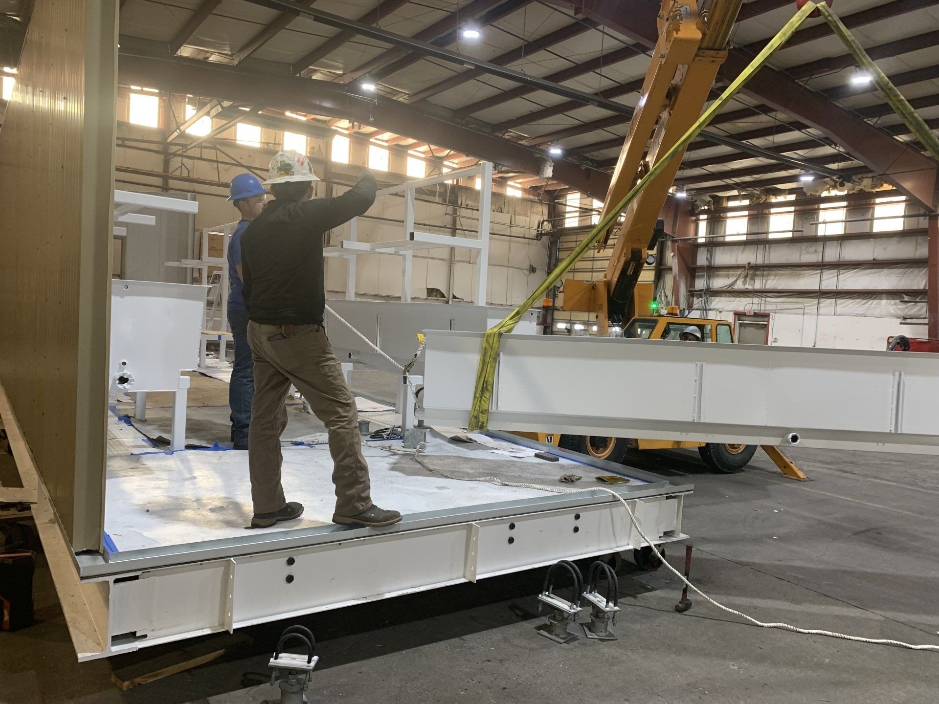 Worker guiding a suspended white panel in a warehouse during assembly