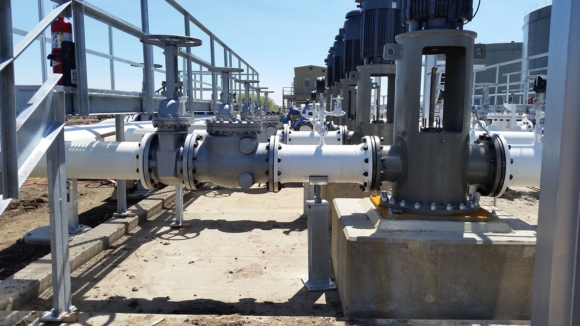 Industrial pipes and valves in a utility plant, with metal railings and concrete supports under a clear sky