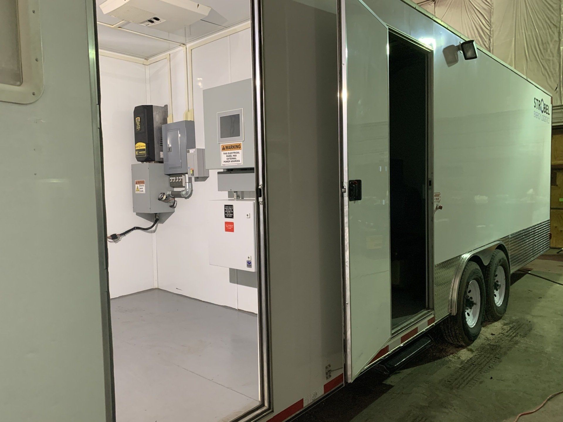 Open trailer doorway showing a small white utility room with electrical panels and wall-mounted equipment