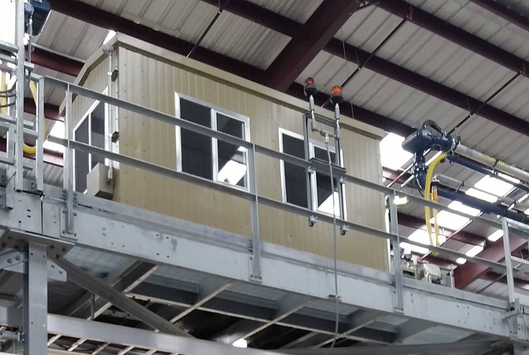 Workers on a raised industrial platform inside a warehouse, surrounded by metal scaffolding and roof beams.