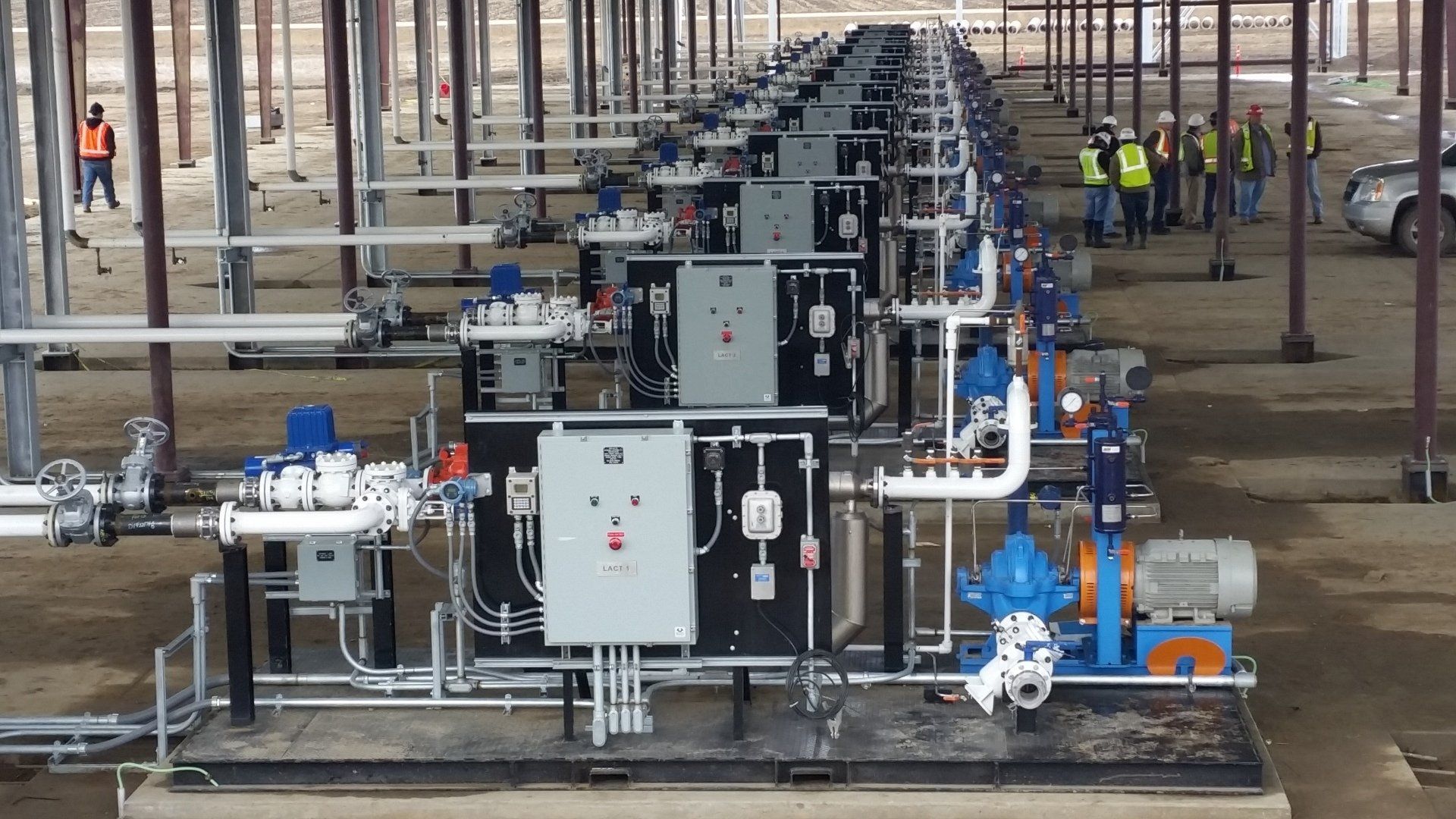 Industrial piping and control units lined up in a large warehouse, with workers in the background