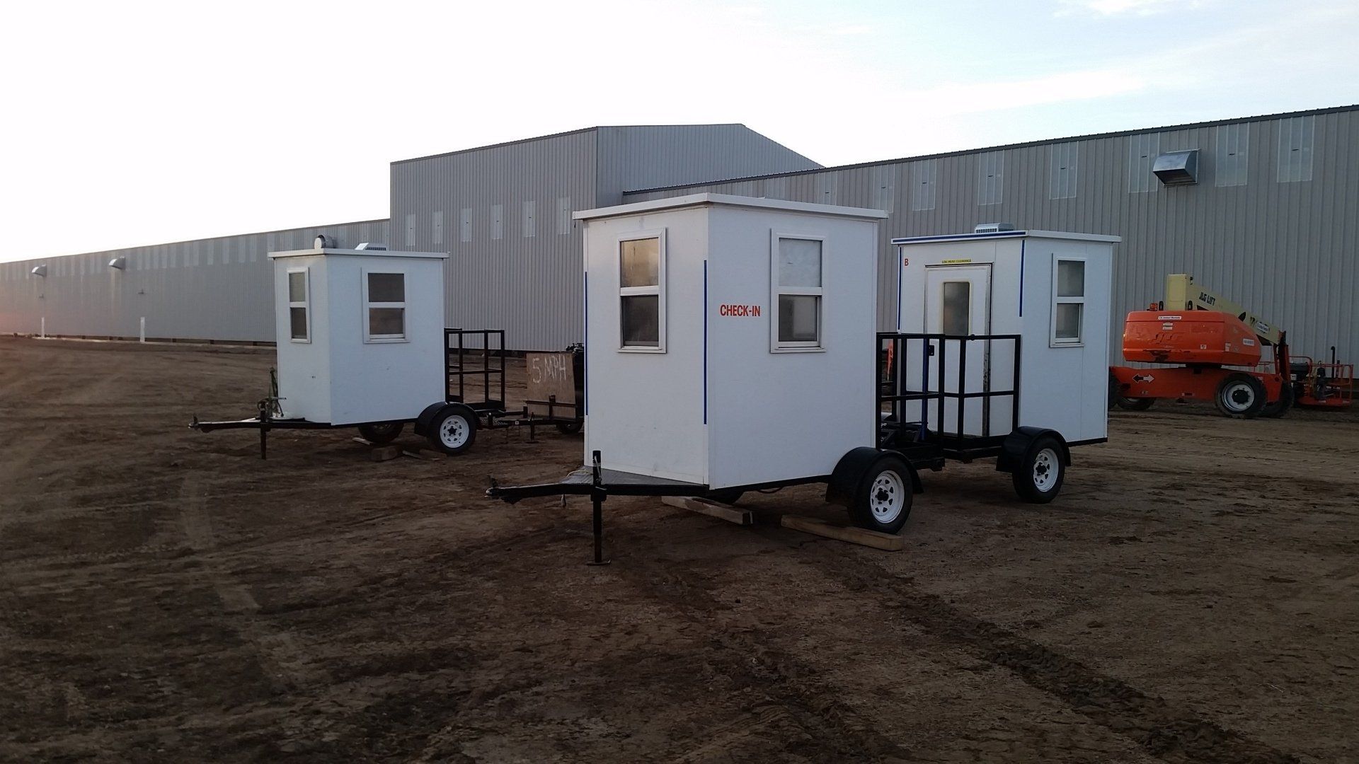 Three small white mobile cabins on trailers in a dirt lot beside a large metal building.