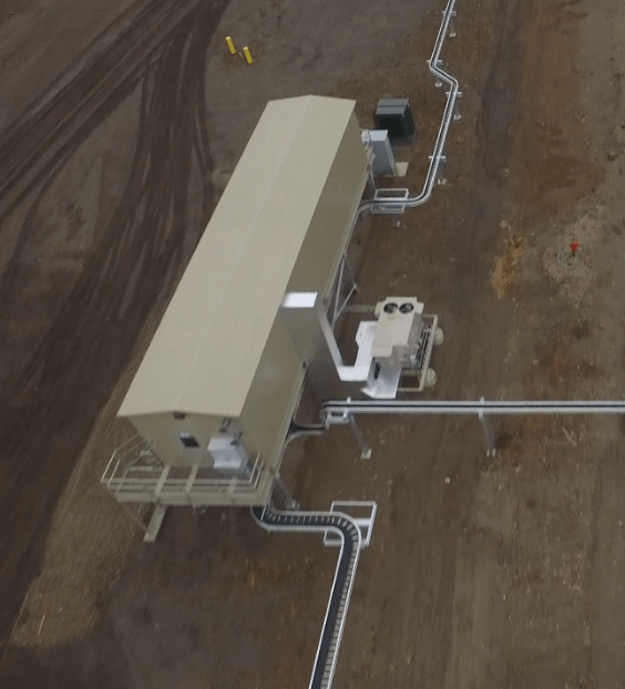 Aerial view of an industrial site with a beige utility building, pipes, and equipment on bare ground.