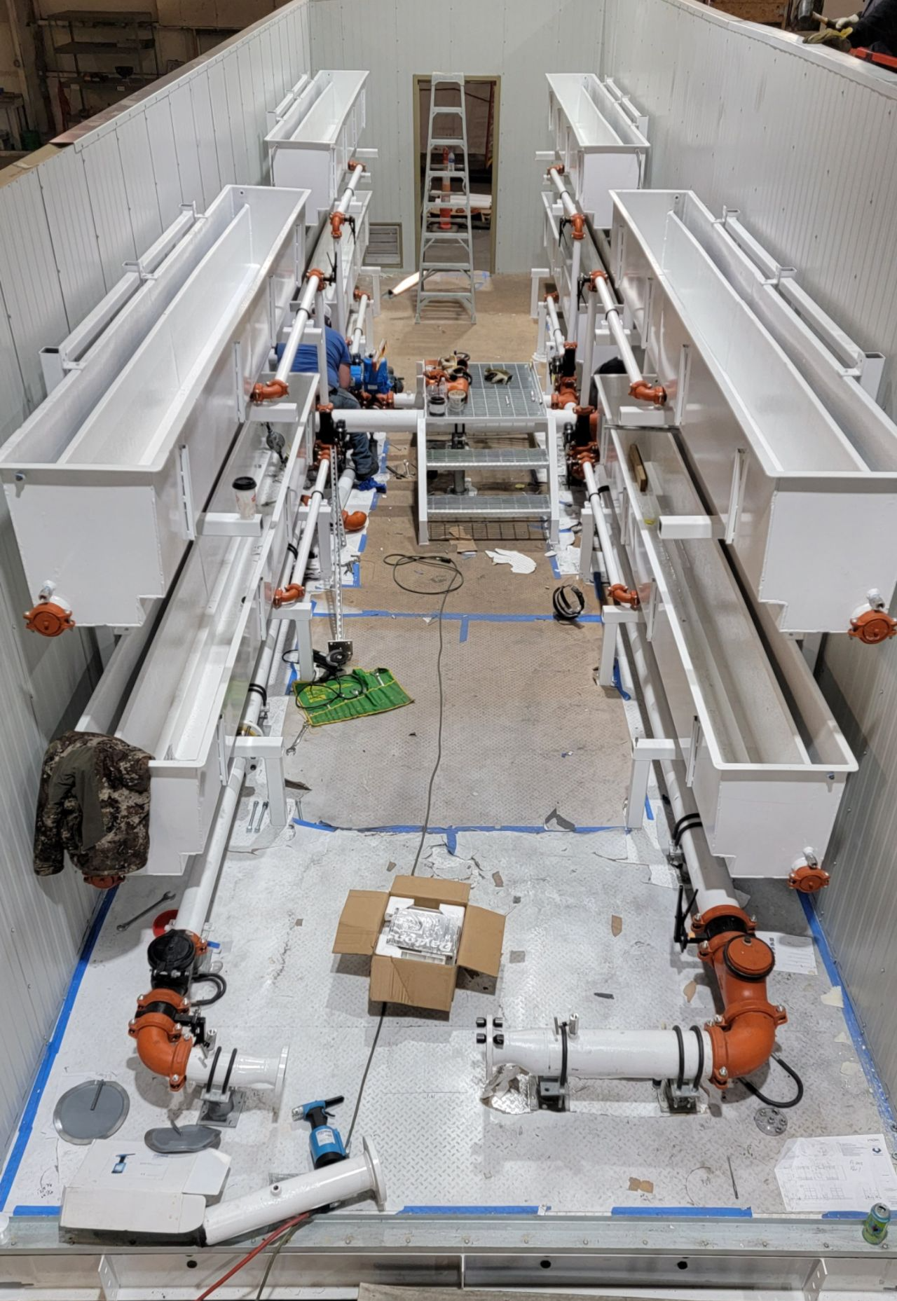Top-down view of a large industrial tunnel with white walls, machinery, and orange equipment on both sides.