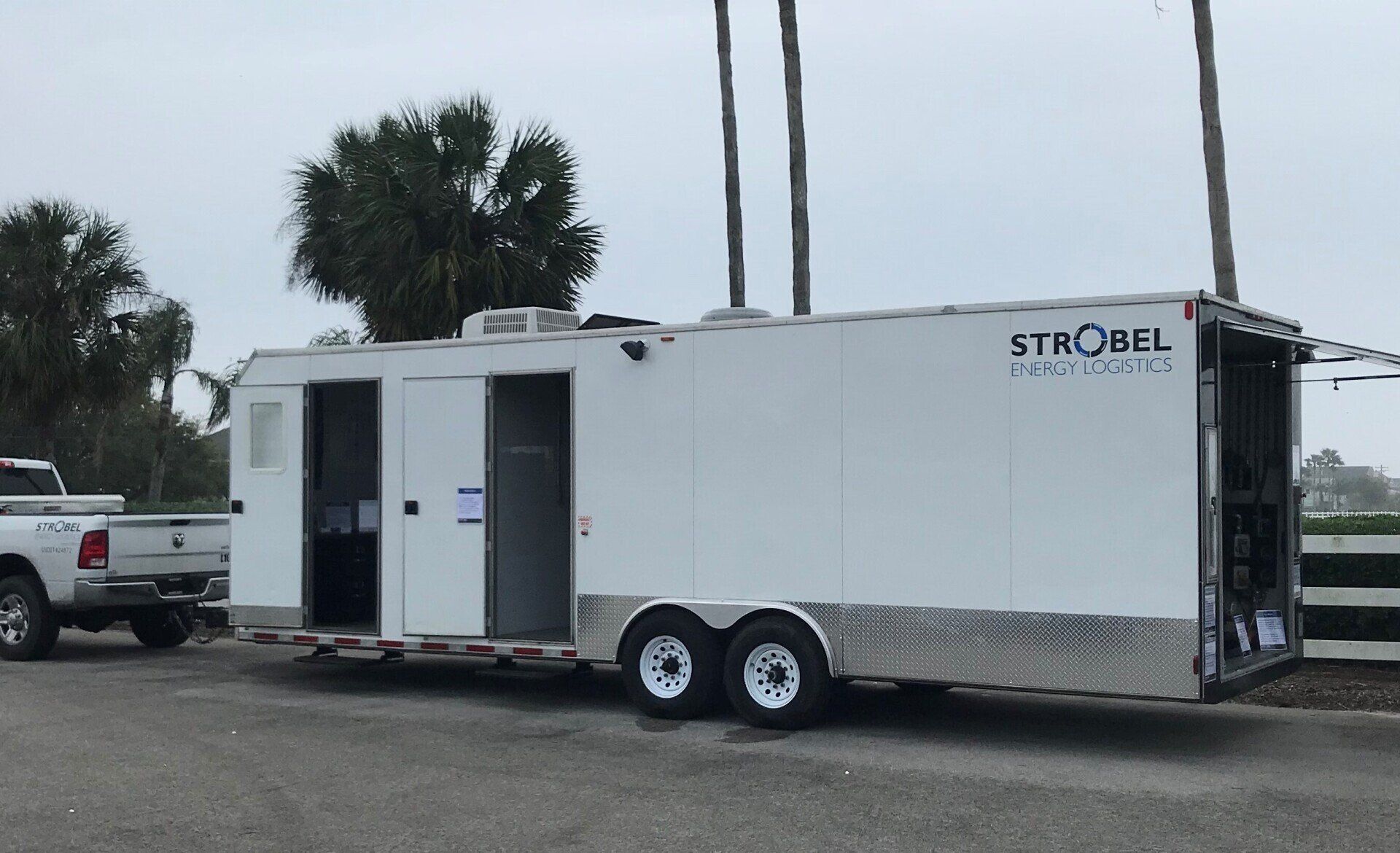 White mobile trailer parked by a pickup truck in a lot, with palm trees in the background.