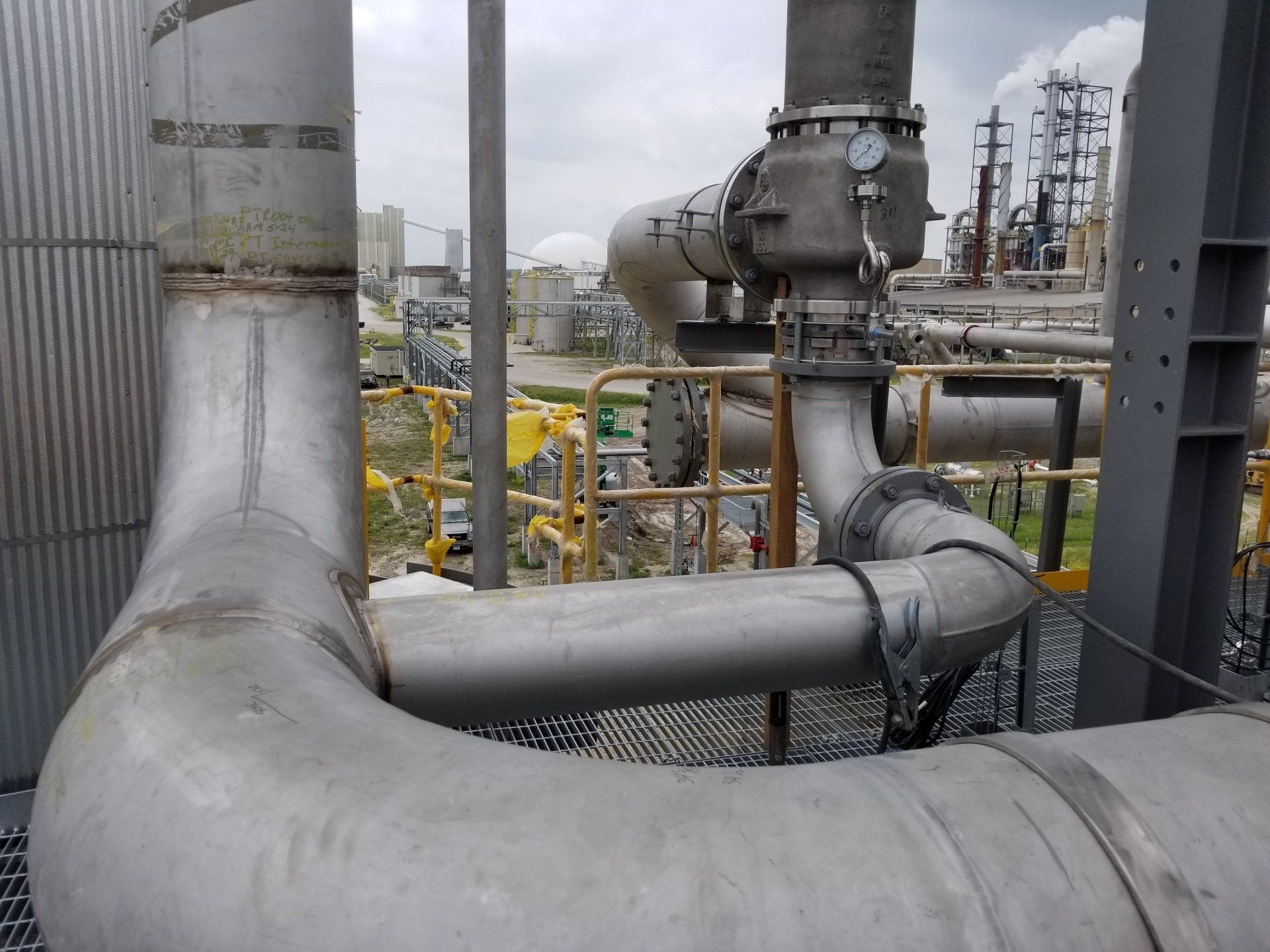 Industrial piping and valves in a refinery setting, with large gray pipes and yellow railings.