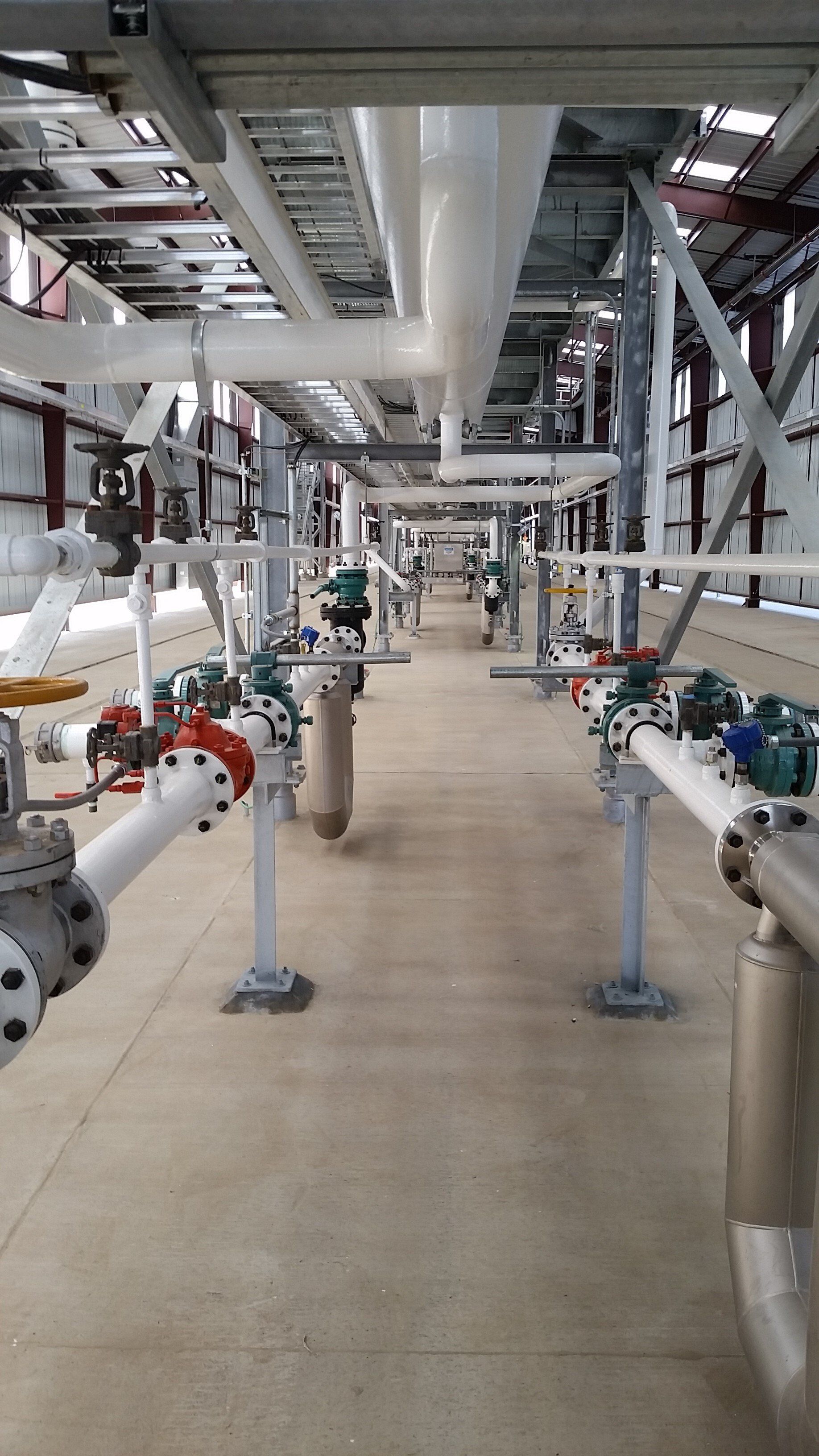 Industrial plant interior with rows of pipes, valves, and overhead steel structures in a clean corridor