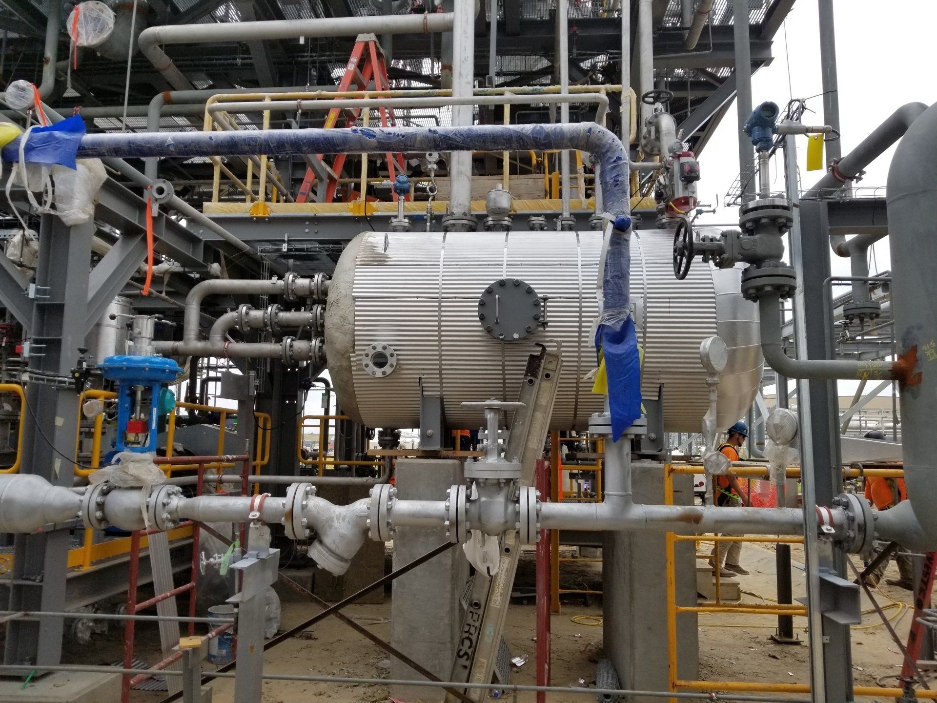 Industrial plant with pipes, tanks, and workers in blue safety gear on a metal platform.
