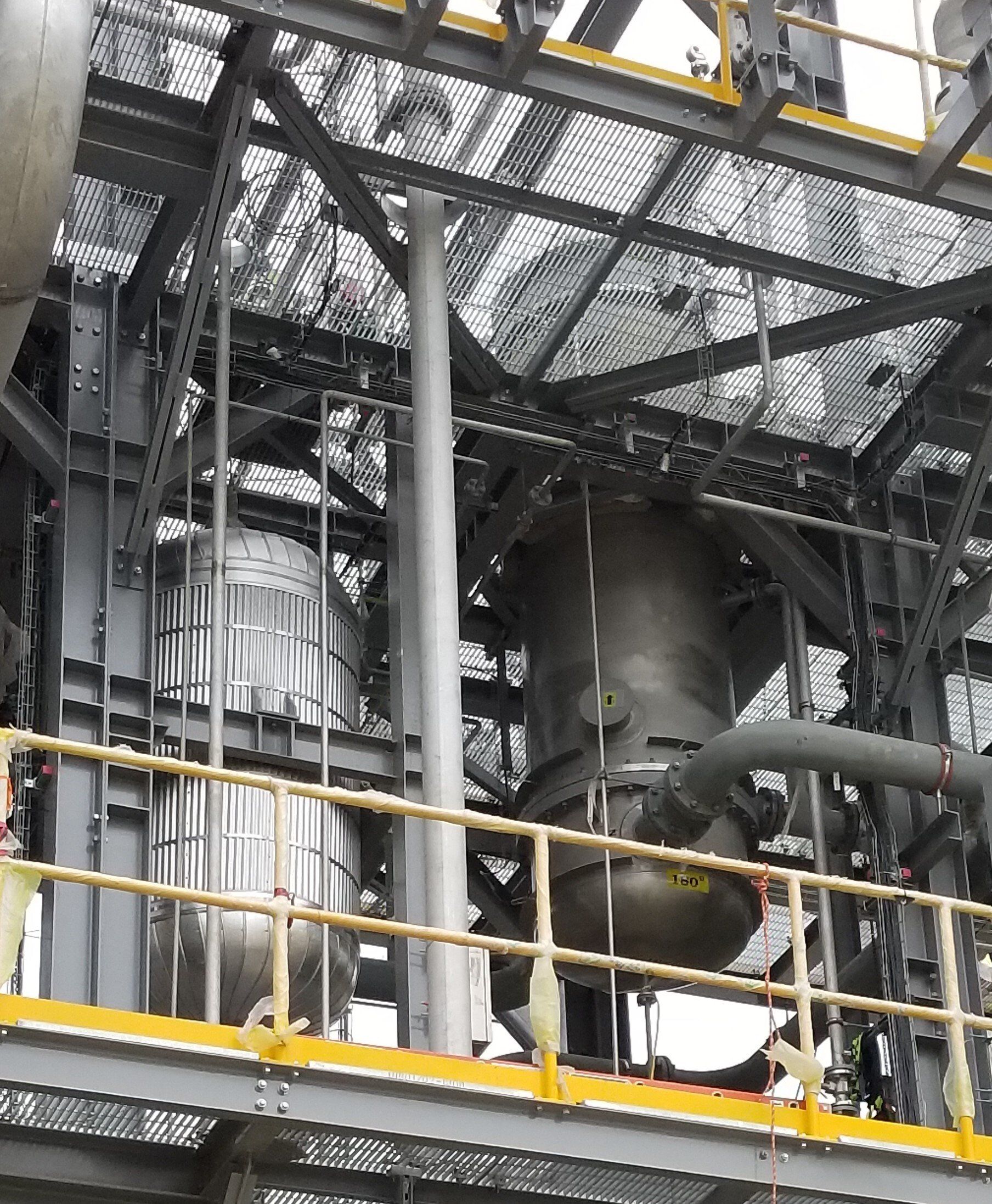 Industrial machinery behind metal railings and grating in a factory setting, with gray pipes and yellow safety rails.
