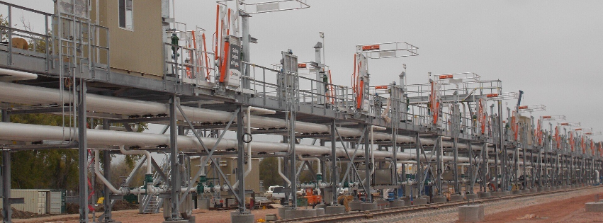 Industrial plant with rows of metal piping, platforms, and cranes in a dusty yard