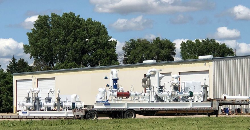 Industrial machinery on a flatbed trailer beside a beige building, with trees and a cloudy sky in the background