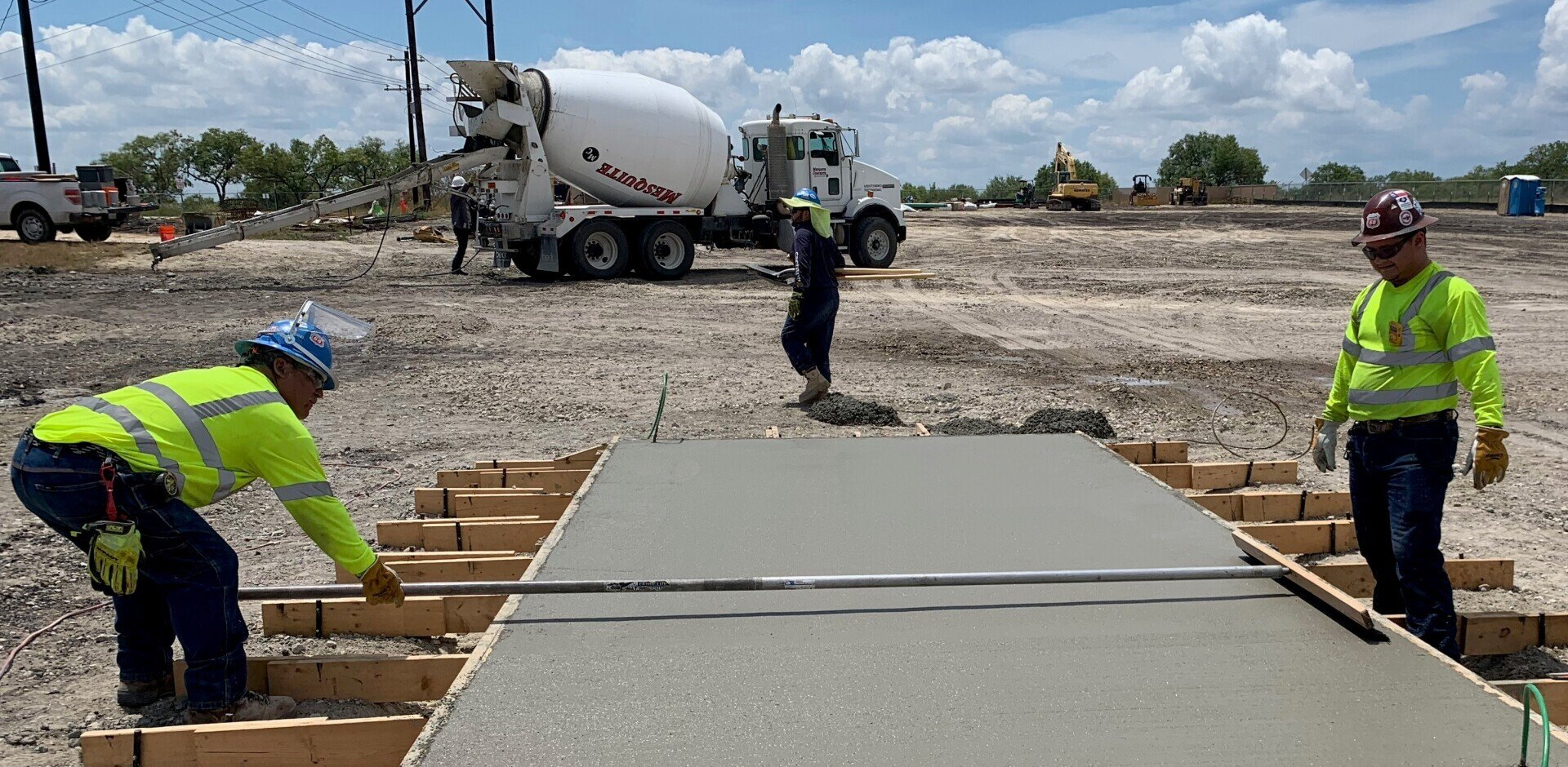 Construction workers smoothing fresh concrete on a roadway form with a cement truck in the background