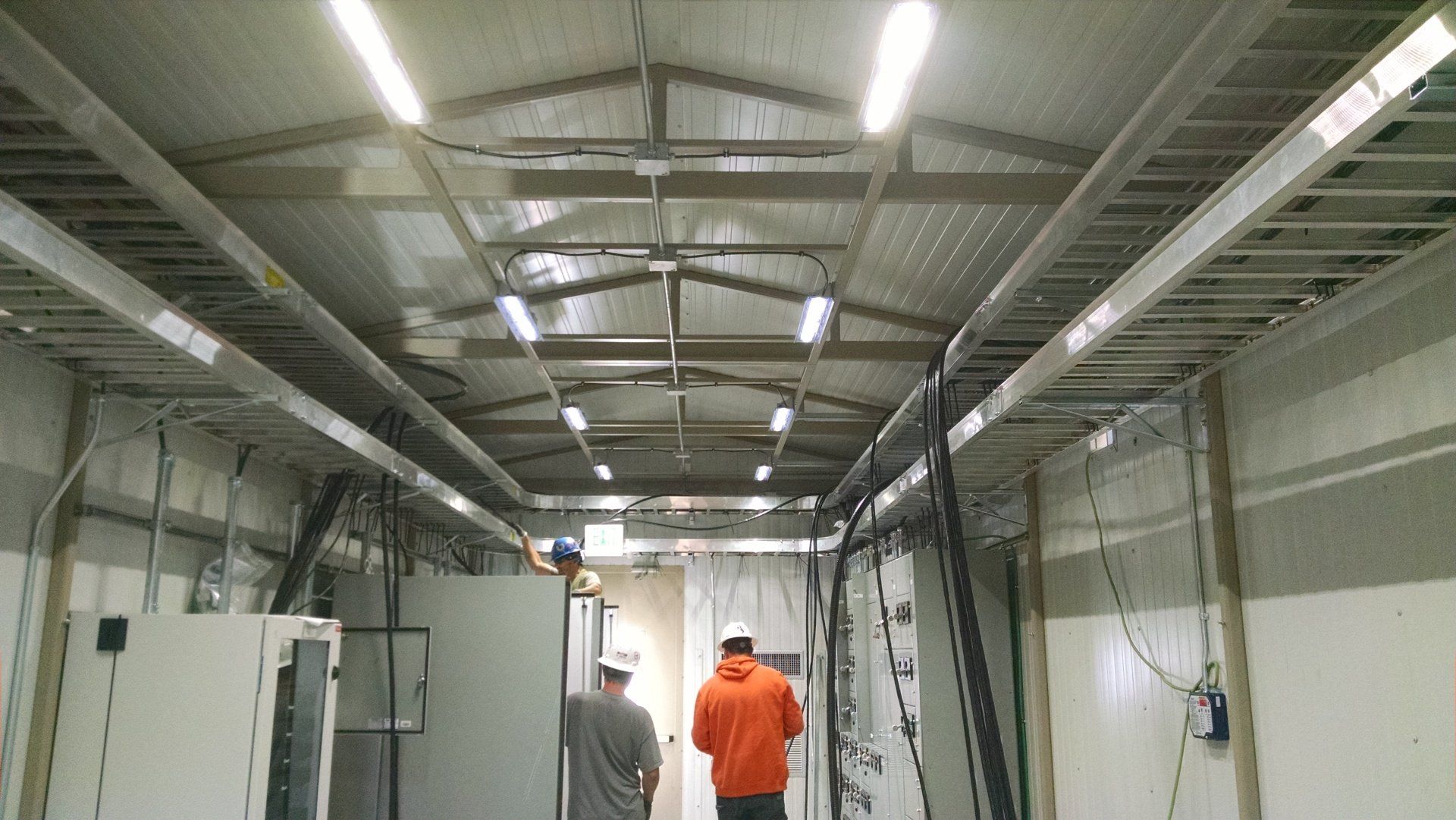 Industrial corridor with exposed ceiling lights and two workers, one in an orange jacket, walking away