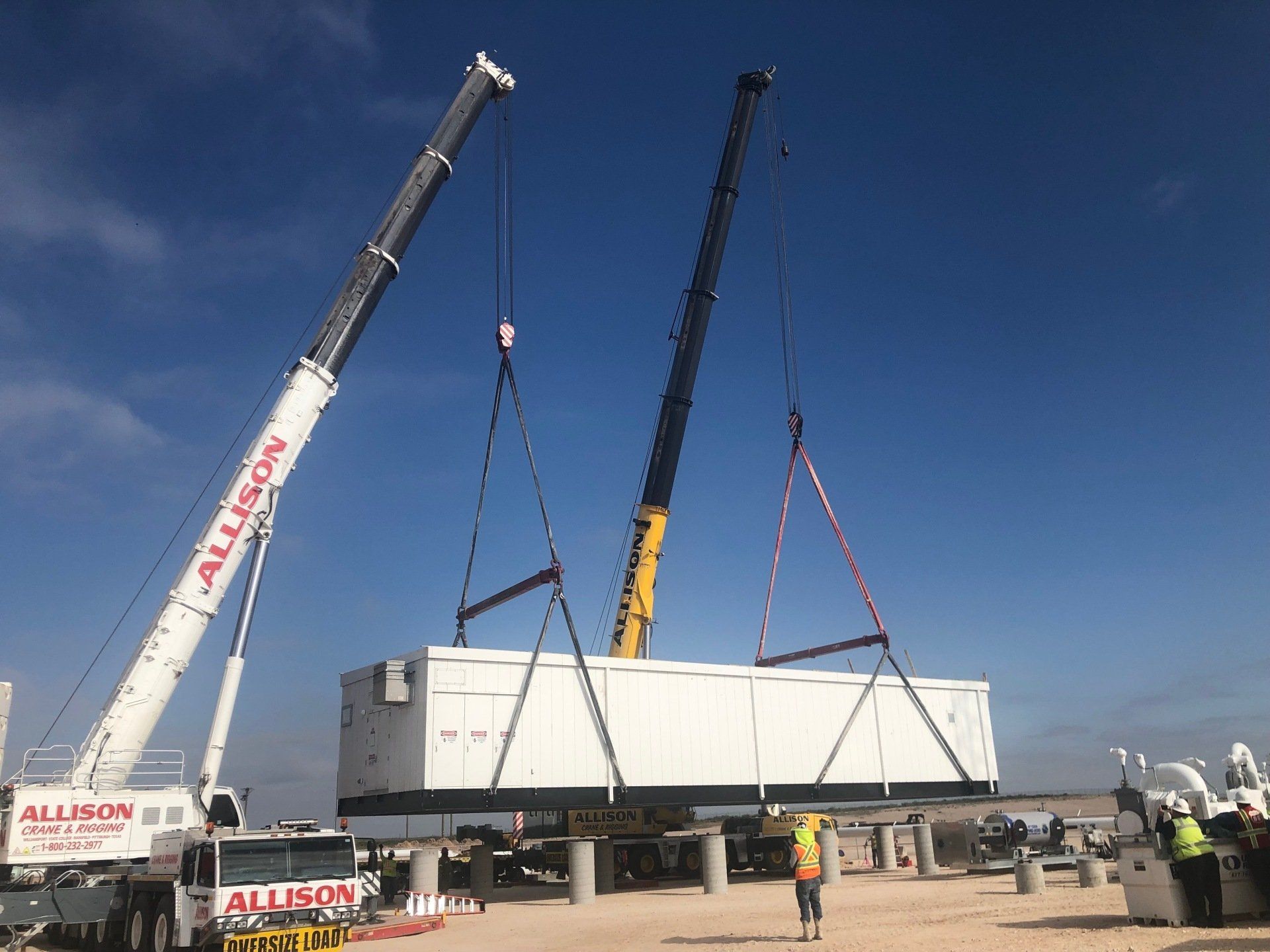 Two cranes lifting a large white modular building at a construction site under a clear blue sky