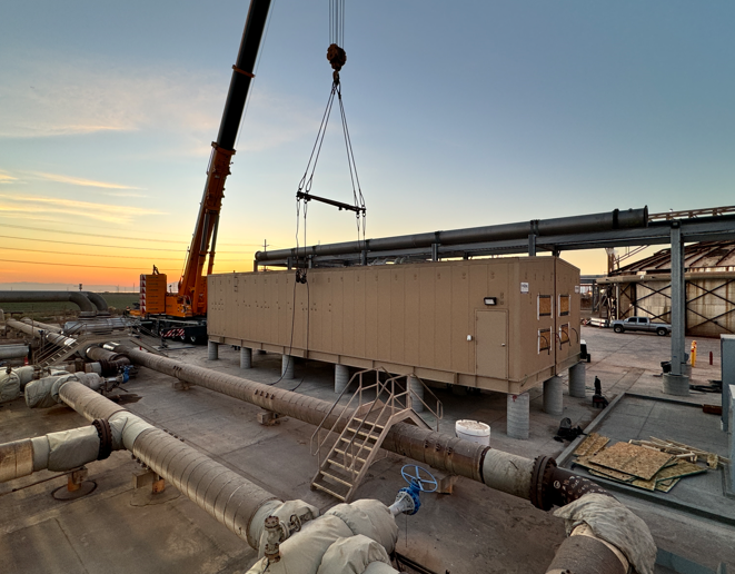 Crane lifting a beige industrial module at an outdoor plant during sunset.