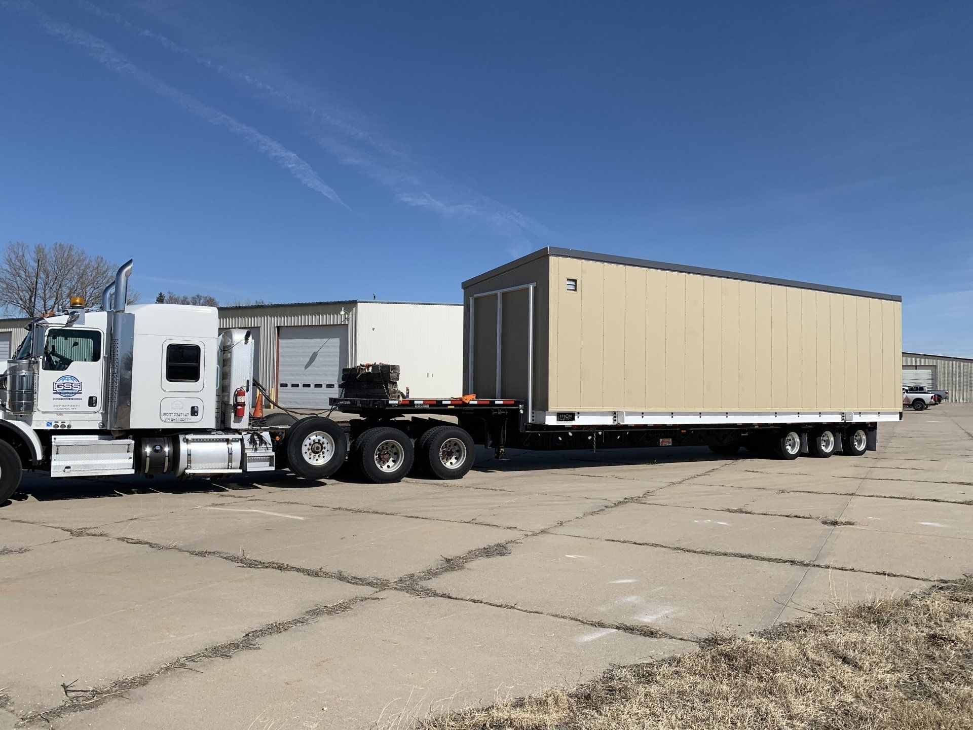 Semi truck hauling a beige trailer parked on a concrete lot under a blue sky