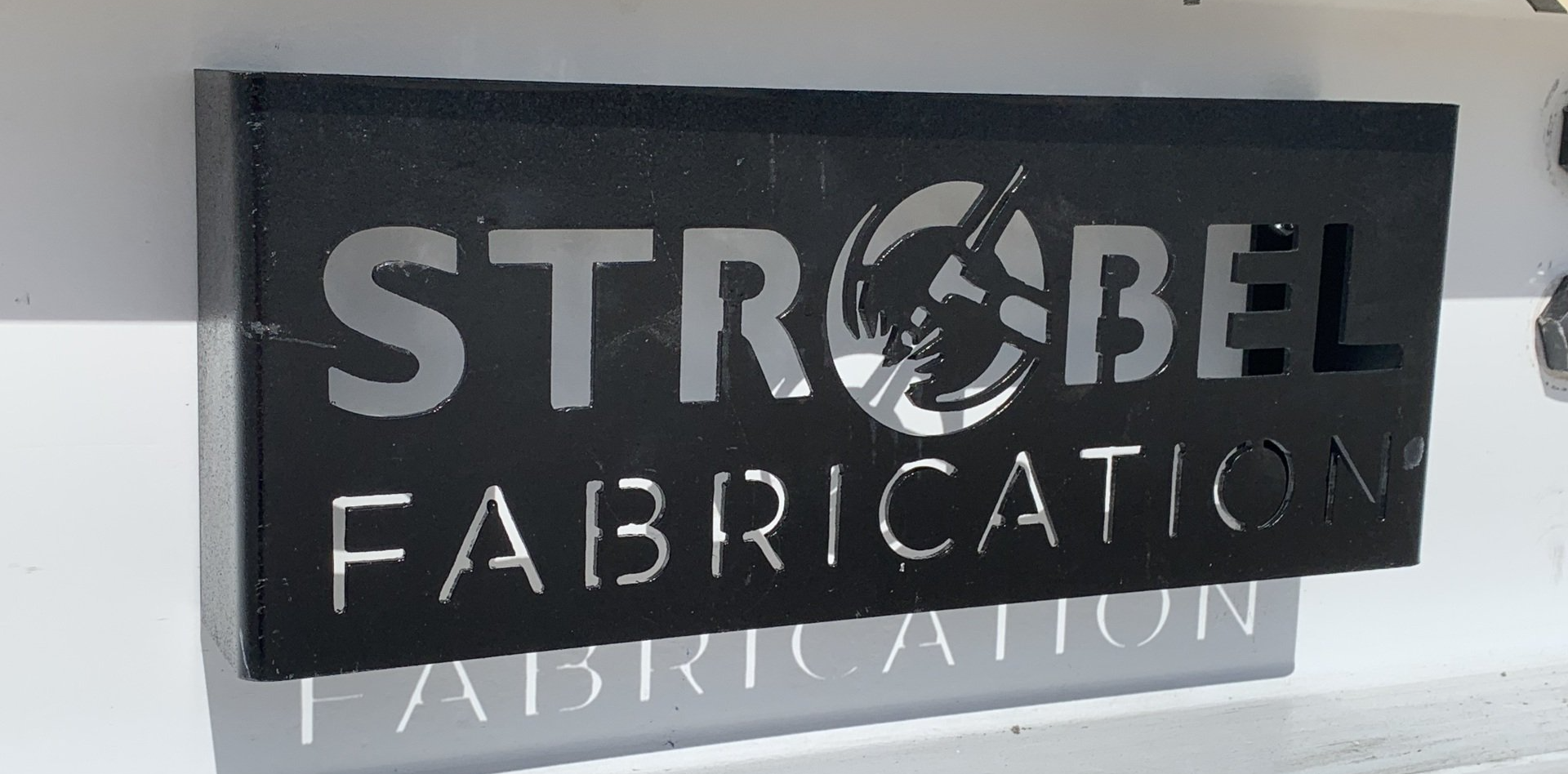 Black sign reading “STROBEL FABRICATION” with a circular logo above a reflective surface.