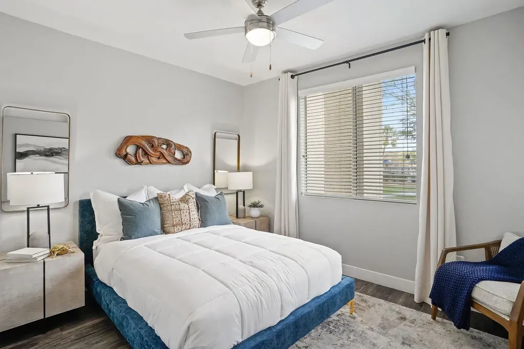 Modern apartment bedroom with a blue upholstered bed, white bedding, two nightstands, and a window with blinds.