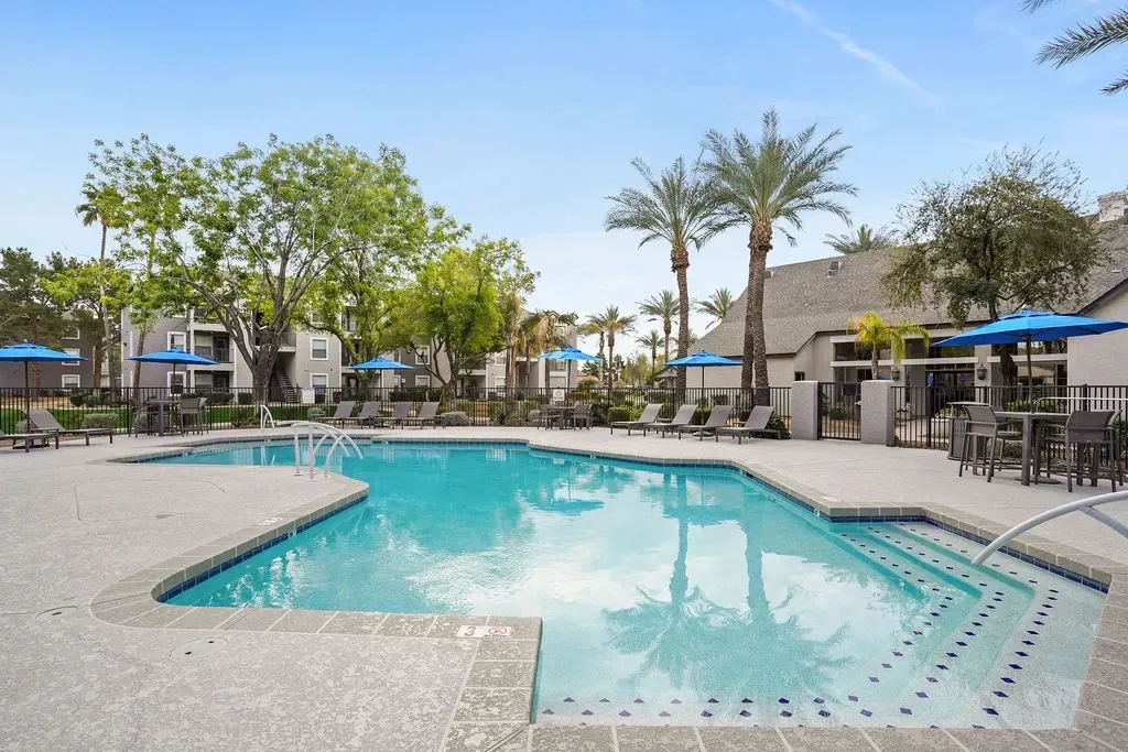 Outdoor apartment pool area with lounge chairs, blue umbrellas, and palm trees.