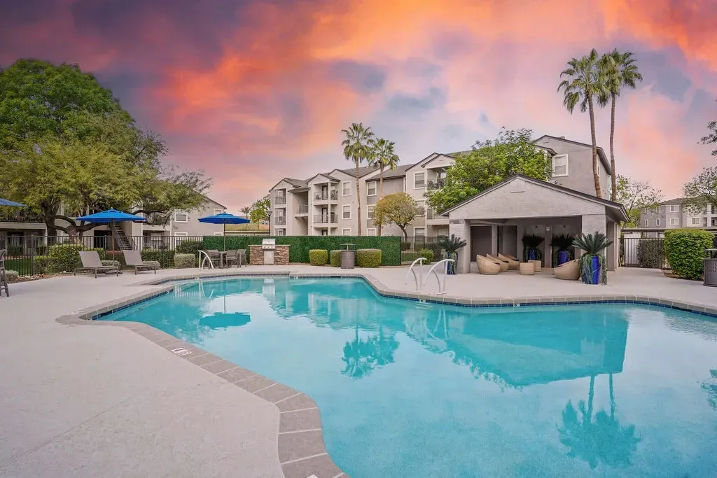 Outdoor apartment community pool with lounge chairs, blue umbrellas, and palm trees.