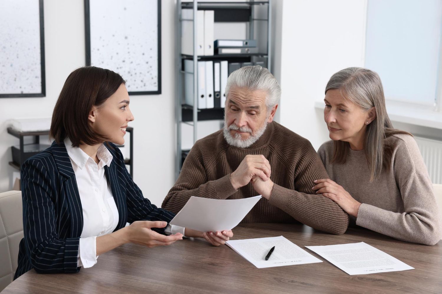 Three people in a meeting review papers at a table in a bright office.