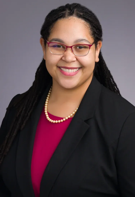 Smiling person in glasses wearing a black blazer and pink top against a gray background