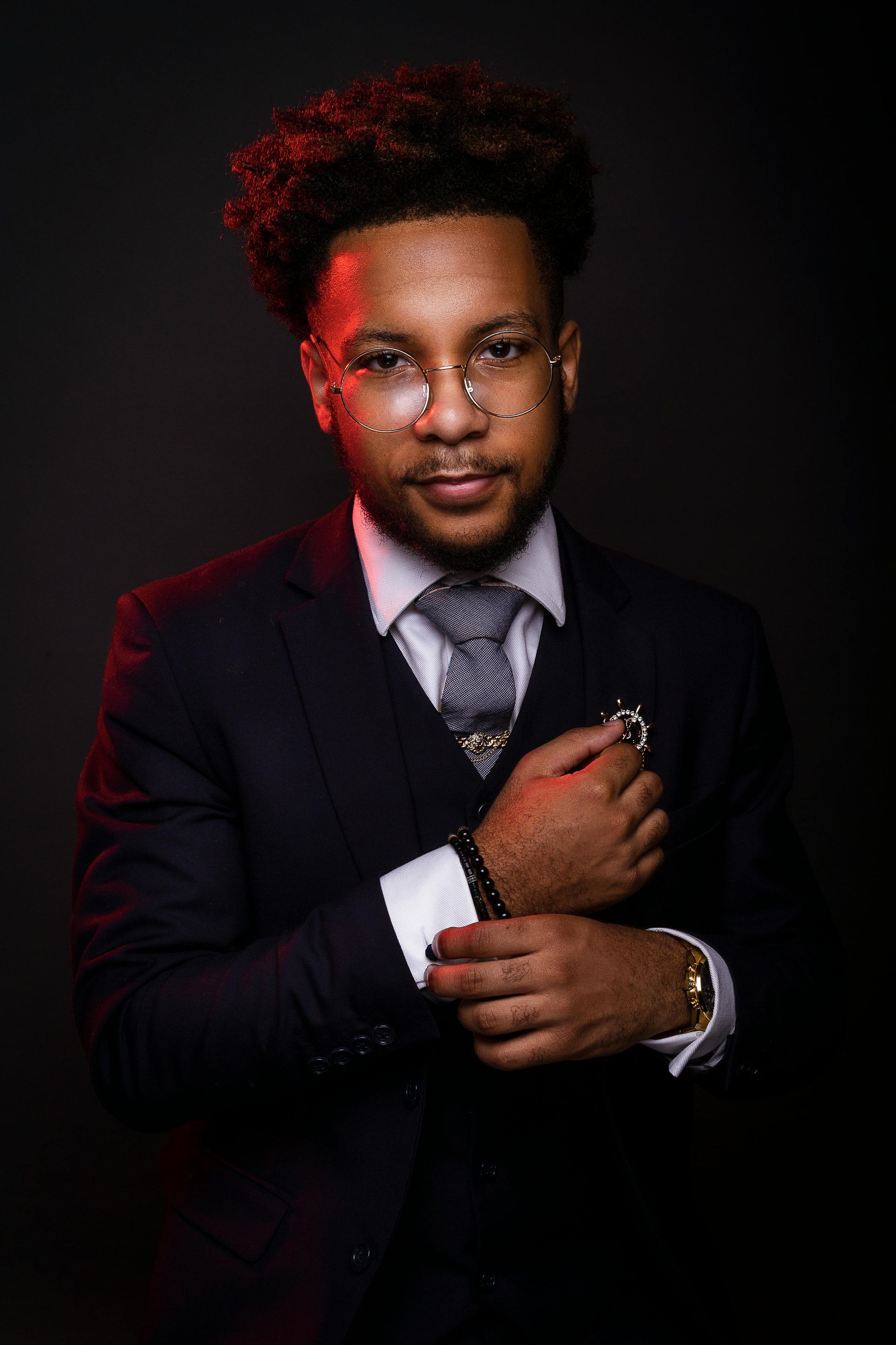 Man in a dark suit adjusting his cuff, wearing glasses and a patterned tie, lit dramatically against a black background.