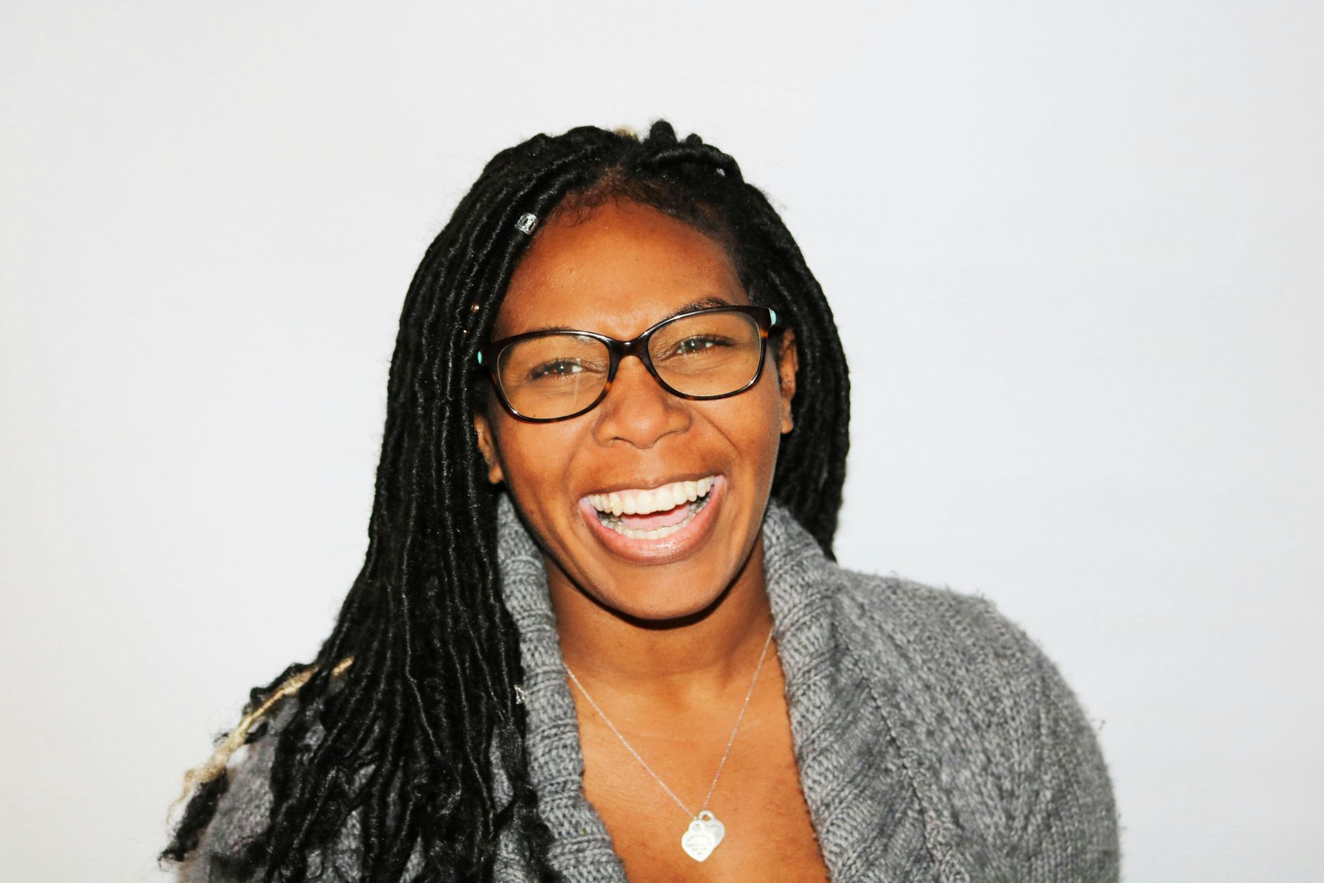 Woman with glasses and long dark braids laughs, wearing a grey sweater in front of a white background.