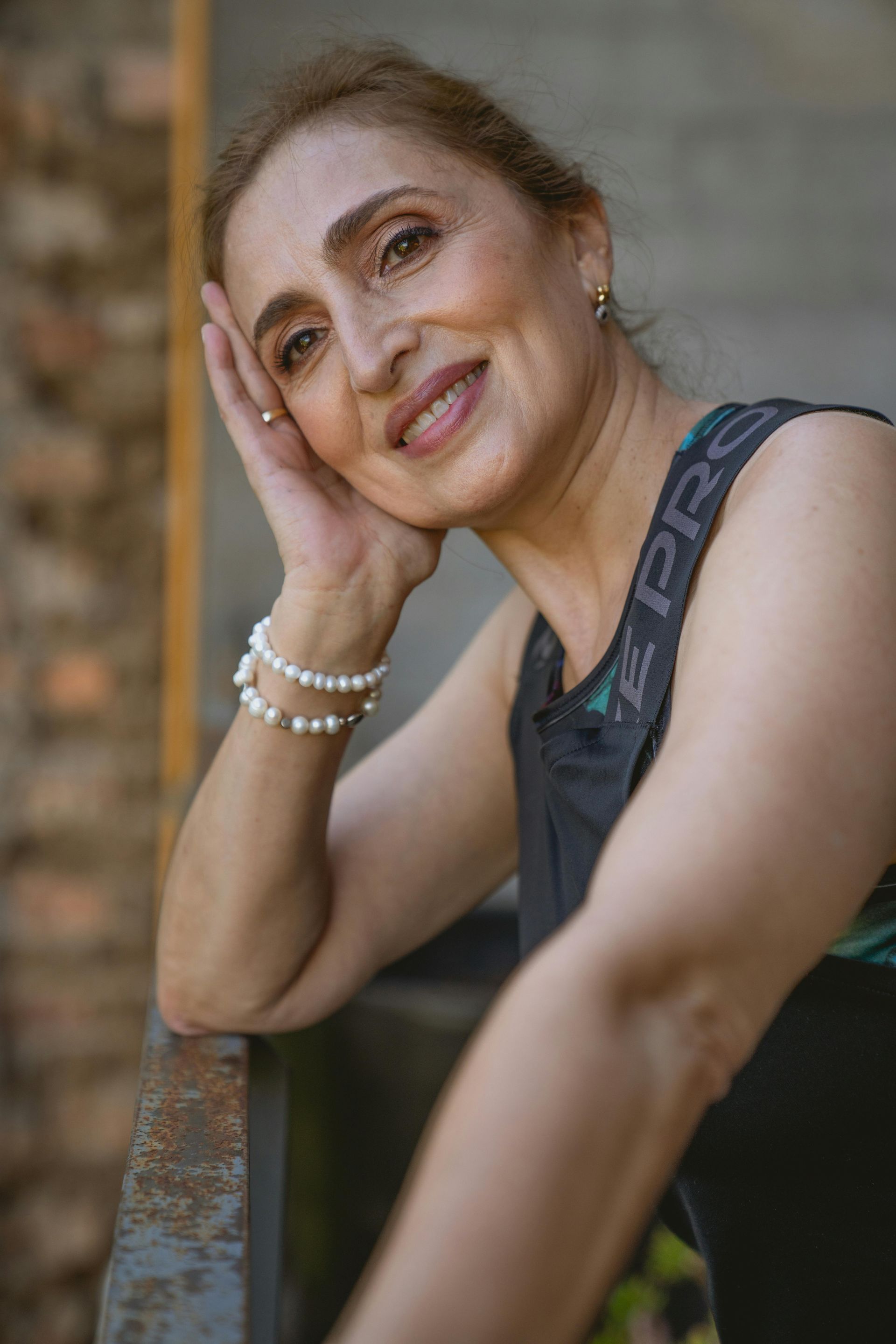 Woman smiling, resting head on hand, wearing a pearl bracelet, leaning against a railing.