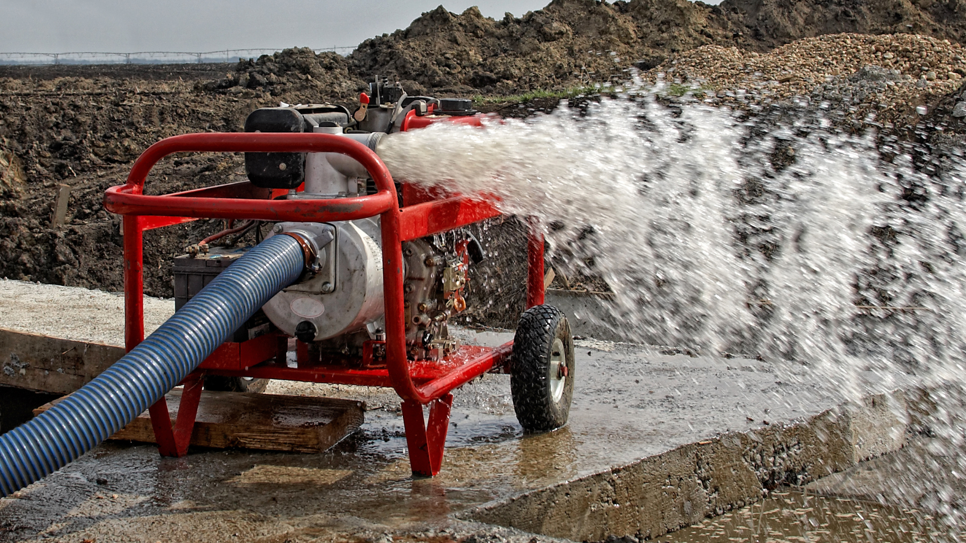 Red water pump discharging water onto a concrete surface, with a backdrop of mud and rocks.