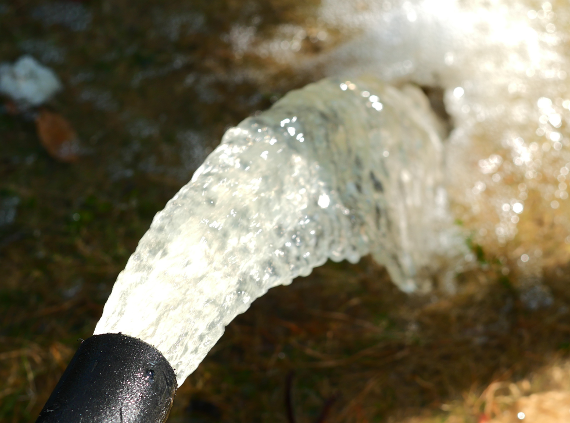 Water gushing from a black hose onto the ground, splashing and creating a foamy texture.