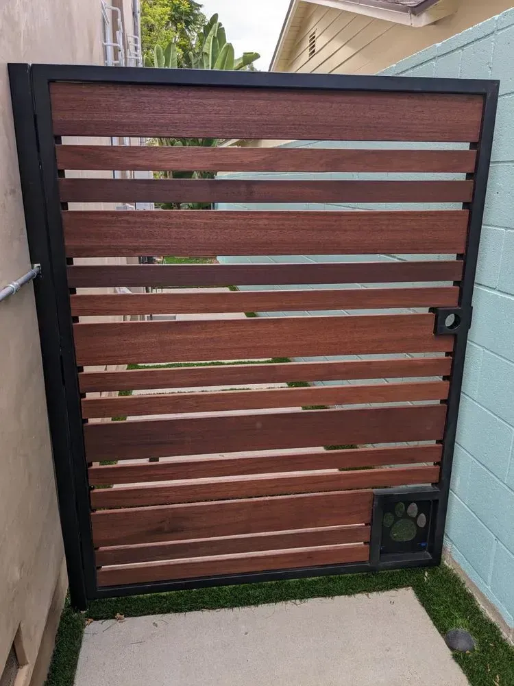 Wooden gate with horizontal slats and a built-in dog door, framed in black metal, next to a teal wall.