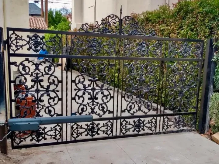 Black wrought iron gate with ornate scrollwork, automated with a motor, set against a green hedge.