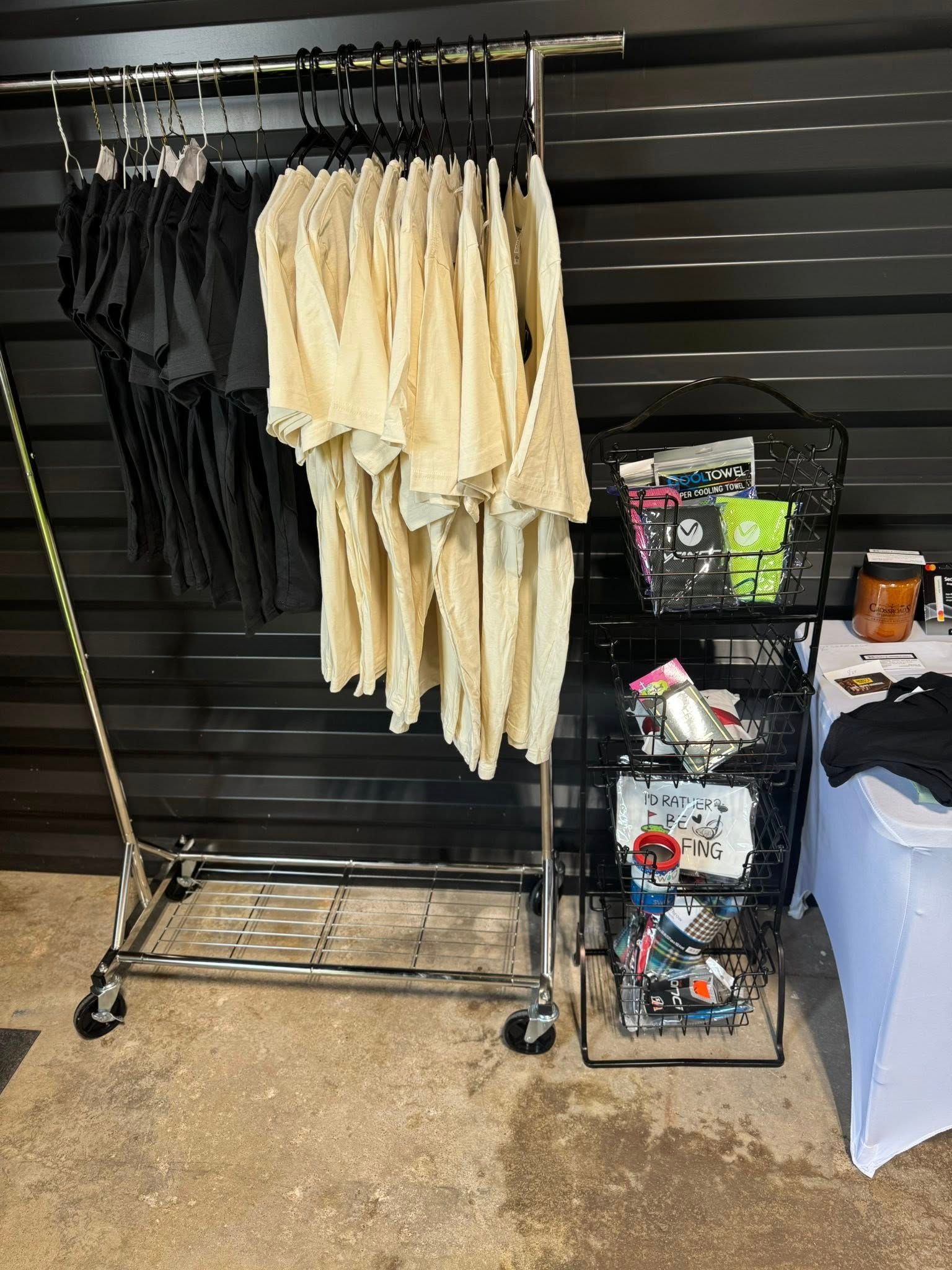 Clothing display: Black shirts, cream tops hanging on a rolling rack, wire basket with accessories on the side.