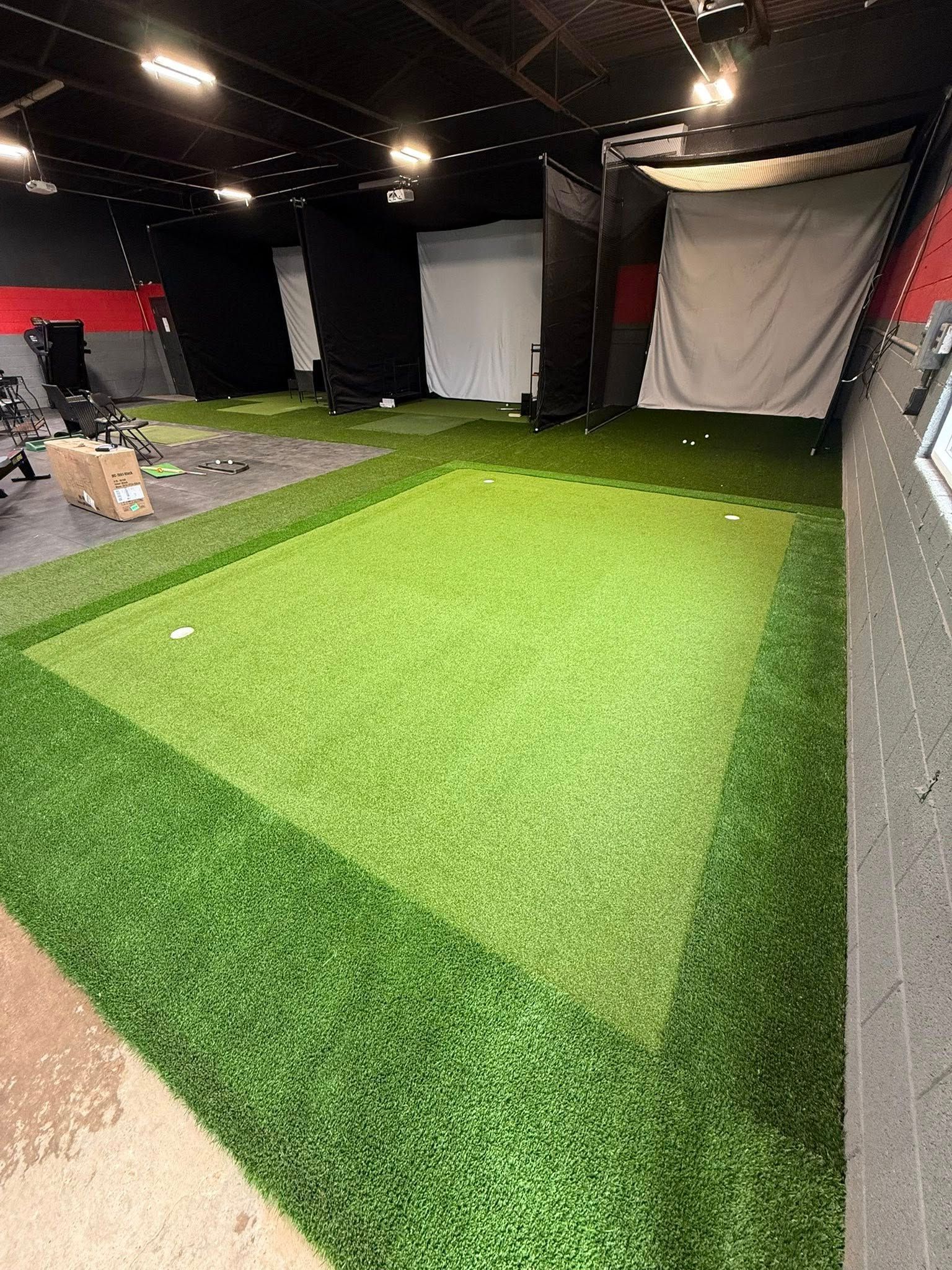 Indoor golf training area with artificial turf, nets, and screens.