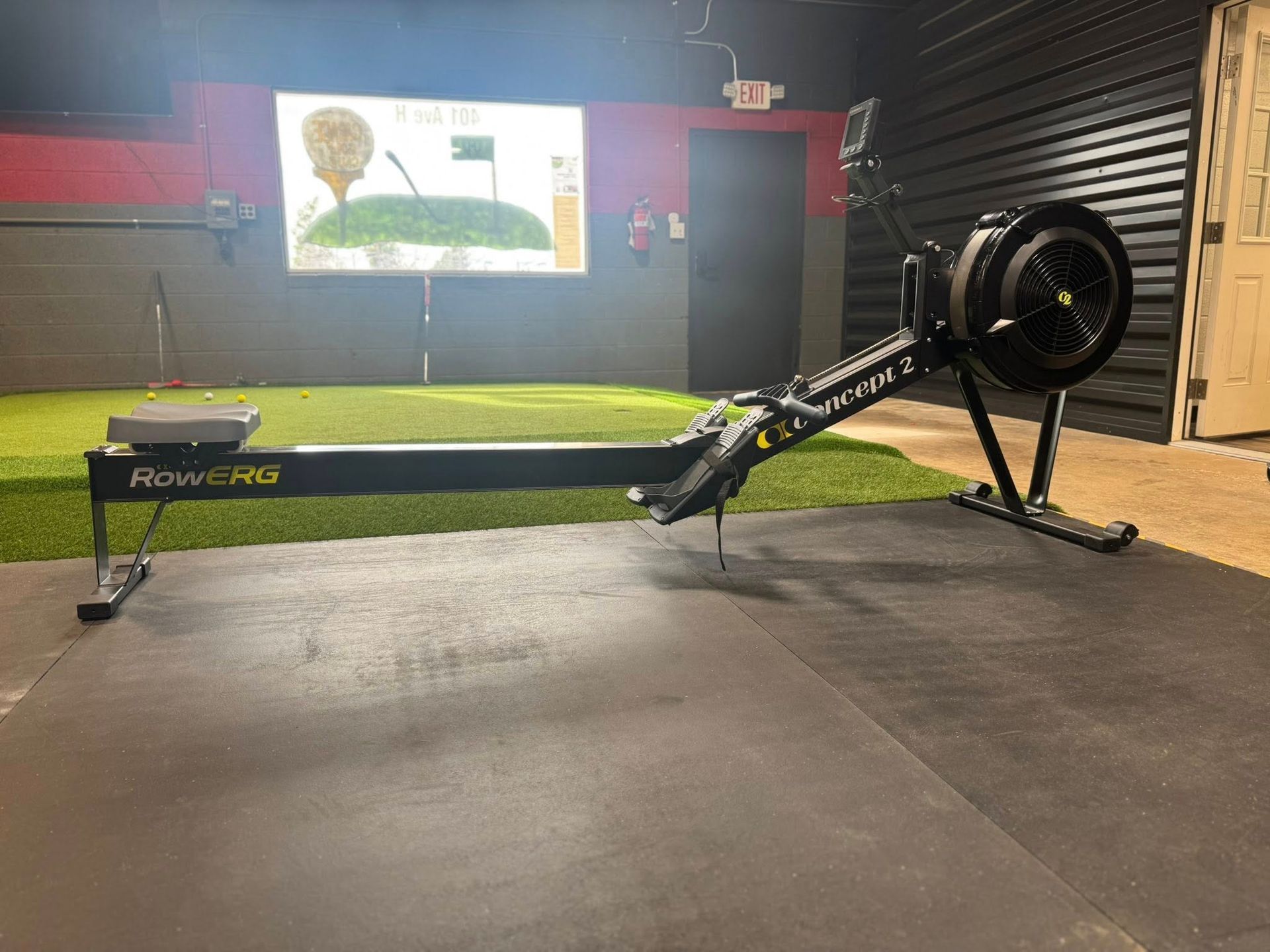 A black rowing machine in a gym setting with a golf simulator projected on the wall.