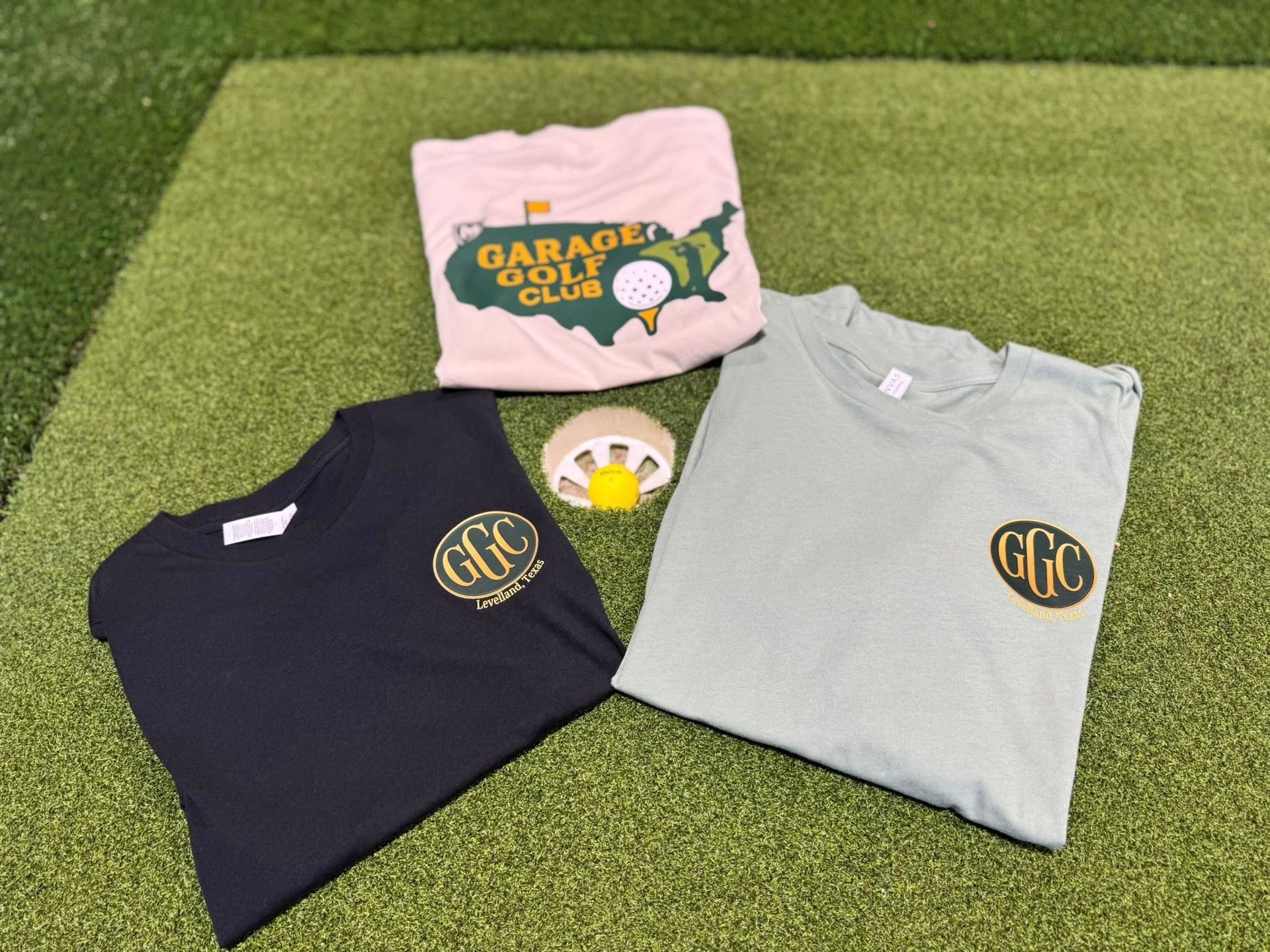 Three t-shirts and a golf ball on artificial turf. One shirt is black, one green, one cream-colored. 
